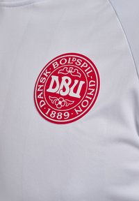 Vit atletisk tröja med en framträdande röd cirkulär logotyp som inkluderar "DANSK BOLDSPIL UNION" och "1889" i stiliserad text.