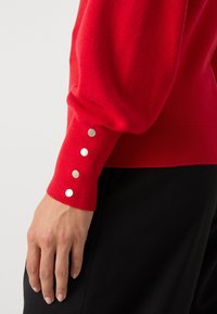 Czerwony sweter z dzianiny o dopasowanym kroju, z bufiastymi rękawami i srebrnymi zatrzaskami na mankietach, uzupełniony fakturowanym czarnym materiałem.