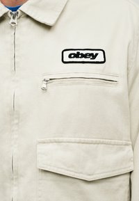 Ελαφρύ μπεζ μπουφάν με φερμουάρ, με κεντημένο λογότυπο "obey" σε πατς, και μια τσέπη στο στήθος με φερμουάρ, καθώς και μια τσέπη με καπάκι από κάτω.