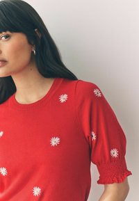 Pull en tricot rouge avec des manches courtes bouffantes, orné de marguerites blanches brodées et de détails de smocks texturés sur les bords des manches.