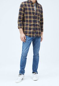 Camicia di flanella a quadri in blu navy e giallo, con bottoni e maniche arrotolate; abbinata a jeans blu e sneakers bianche. Design semplice, vestibilità casual.