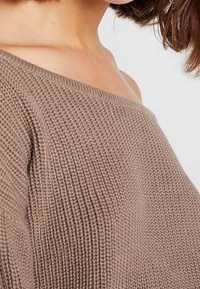 Pletený svetr s odhalenými rameny v teplé taupe barvě, se žebrovanou texturou a volným střihem, ukazující jemné, pohodlné detaily látky.