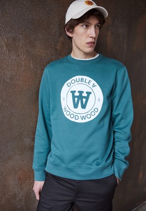 Teal sweatshirt with a round logo displaying "DOUBLE A WOOD WOOD" in white and dark green. Paired with a beige cap. Smooth texture.