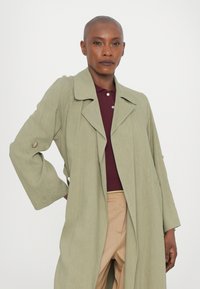 Cappotto lungo verde chiaro con maniche ampie, dettagli in bottoni e colletto con alette, indossato sopra un polo bordeaux e pantaloni beige.