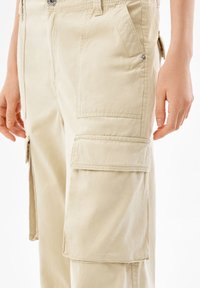 Beige cargo-housut, jotka on valmistettu puuvillasta ja joissa on useita sivutaskuja, nappisuljenta ja näkyvät saumat. Sileä pinta ja rento leikkaus.