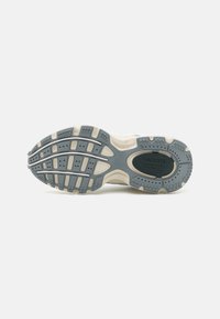 Semelle d'une chaussure Lacoste avec une bande de roulement en caoutchouc gris et beige, grip texturé, et le nom de la marque ainsi que le logo embossés au niveau du talon.