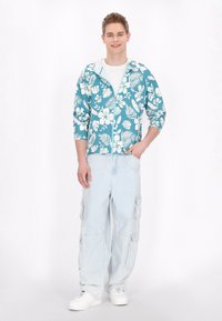 Ljust blå blommig zip-up hoodie med vita hibiskus- och lövmönster, parat med ljus denimcargo byxor och vita sneakers.