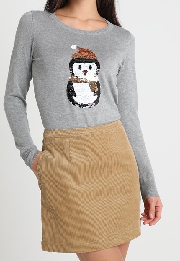 Femme portant un pull gris avec un manchot orné de sequins et un bonnet de Père Noël, ainsi qu'une jupe en velours côtelé beige avec des poches.