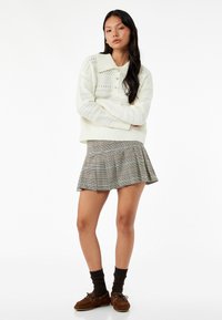 Weißer Strickpullover mit einem Kragen, kombiniert mit einem grauen karierten Faltenrock. Braune Wildlederschuhe und dunkle Socken runden den Look ab.
