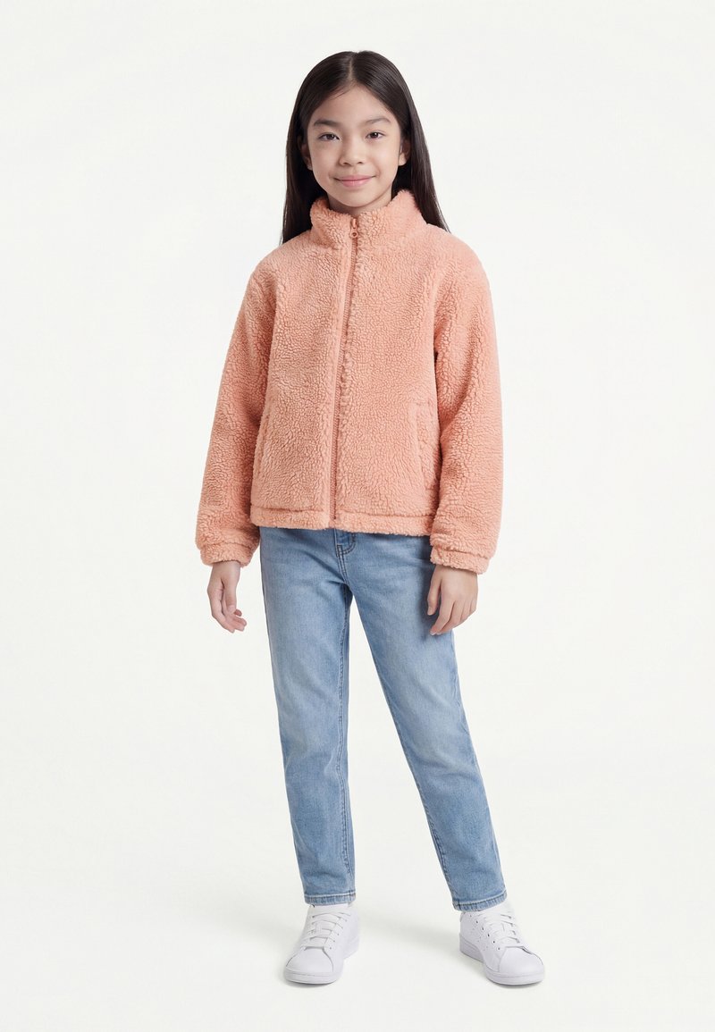 Ragazza in piedi che indossa una giacca in pile color pesca, jeans azzurri chiari e sneakers bianche su sfondo bianco uniforme.