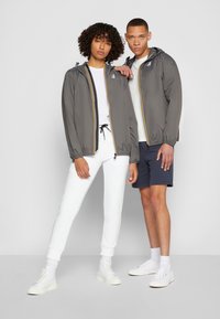 Deux mannequins portent des vestes grises assorties avec des détails en rayures jaunes. Un mannequin en pantalons blancs, l'autre en shorts bleu marine et baskets blanches.