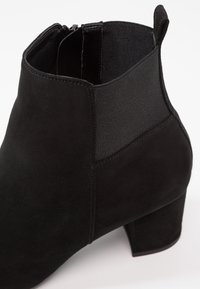 Bottine en daim noire avec talon bloc, panneau latéral élastique, fermeture éclair et languette à l'arrière.