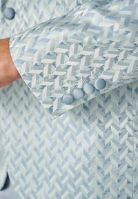 Blazer azzurro chiaro con motivo a spina di pesce. Tessuto morbido con una leggera lucentezza. Manica decorata con quattro bottoni rivestiti in tessuto abbinato.