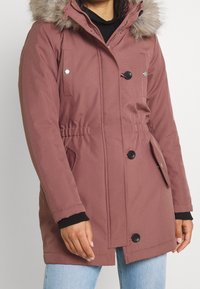 Veste parka marron avec capuche doublée de fausse fourrure. Comprend des boutons noirs, des poches latérales et une taille cintrée pour un look ajusté. Texture lisse.