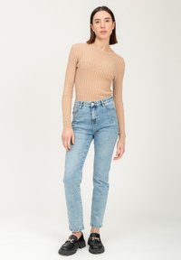Beige geripptes Langarmoberteil, hellblaue hochtaillierte Jeans, schwarze Loafers mit Kettendetail, stehend vor einem neutralen Hintergrund.