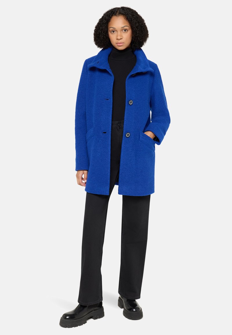 Helblau wollen coat met een kraag, sluiting met knopen aan de voorkant, twee zijzakken en een ontspannen pasvorm, gecombineerd met zwarte broek en stevige laarzen.