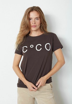 T-shirt en coton marron à manches courtes avec le texte graphique blanc "C°CO" sur le devant. Le modèle tient le bas du t-shirt.