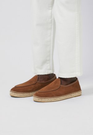 Espadrilles de ante marrón sin cordones con suelas envueltas en yute, combinadas con pantalones blancos recortados sobre un fondo liso.
