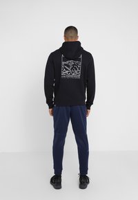 Felpa nera con grafica di montagna bianca e testo "ALASKA STATI UNITI". Indossata con pantaloni tuta blu scuro e scarpe nere, con cappuccio regolabile.