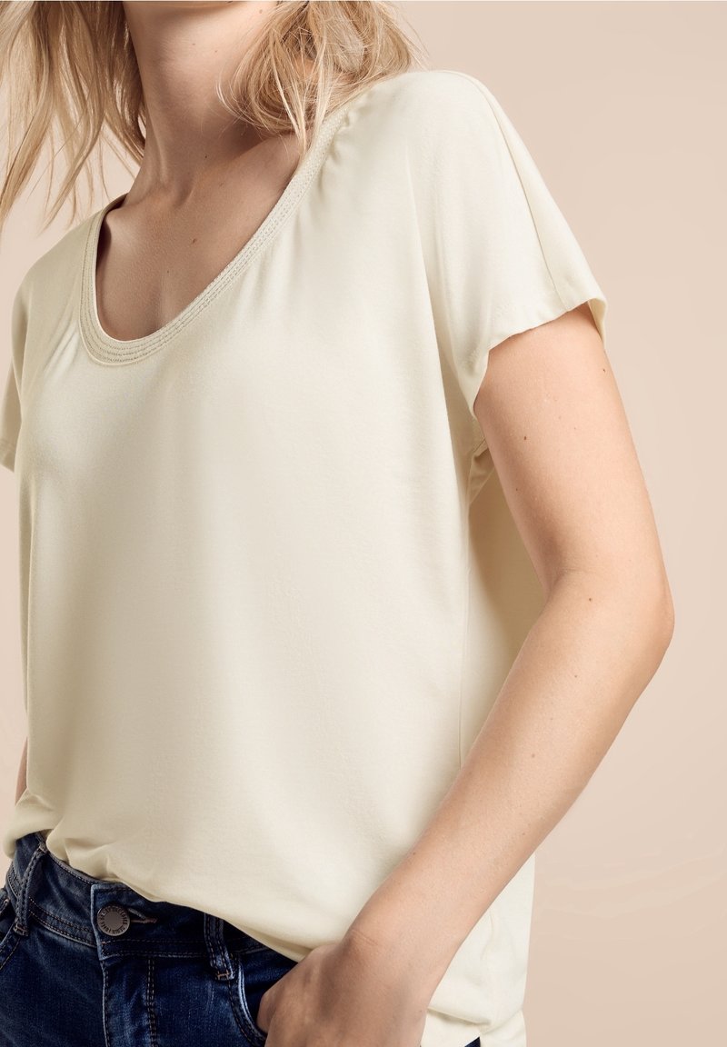 Femme portant un haut crème à manches courtes en V, rentré dans un jean bleu foncé, sur un fond beige.