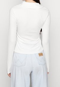 Maglietta bianca a maniche lunghe con collo, abbinata a pantaloni a vita alta di colore blu chiaro con una patch in pelle del marchio e cuciture a zig zag sulle tasche.