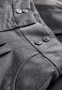 Grijze stof met een gestructureerde weving, met twee zwarte knopen en geplooide details. Het kledingstuk heeft een glad, contrasterend zwart paneel eronder.