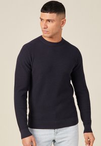 T tumma laivastonsininen ribbipaita, jossa on pyöreä kaula-aukko, pitkät hihat ja kapea helma. Yhdistetty vaaleansinisiin farkkuihin, korostaen rentoa tyyliä.