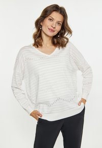 Witte gebreide trui met een V-hals, voorzien van verticale strepen en openwerkdetails. Lange mouwen en een ontspannen pasvorm. Gecombineerd met donkere broek.