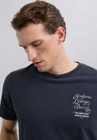 Joven con el cabello corto y marrón mirando hacia abajo, vistiendo una camiseta azul marino con texto blanco y un gráfico de calavera en el pecho.