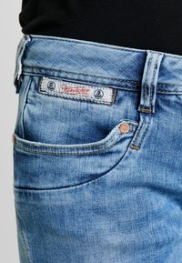 Ljusblå denimjeans med en urtvättad textur. Har en vit etikett med röd skrift, kopparfärgad hårdvara och en klassisk femficksdesign.