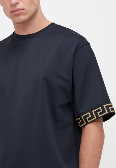 Melna īsām piedurknēm T-kreklu ar teksturētu zelta rakstu apmalēm uz piedurknēm. Izgatavots no gludas, vieglas vielas.