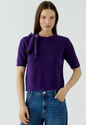 Frau mit glatten braunen Haaren trägt einen lila kurzärmeligen Pullover mit einer Schleife auf der Schulter und blaue Jeans, steht vor weißem Hintergrund.