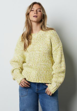 Žena nosi hrubý pletený žltý a biely sveter s rebrovanými manžetami a lemom, v kombinácii s modrými džínami, stojaca pred jednofarebným pozadím.