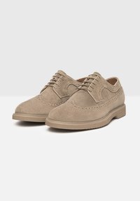 Chaussures en daim beige à lacets avec un design brogue classique, présentant des détails perforés et une semelle en caoutchouc pour une meilleure adhérence.