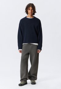 Ung person med lockigt hår som bär en marinblå tröja, vit undertröja, lösa grå byxor och gröna sneakers som står mot en vit bakgrund.