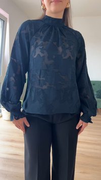Blusa transparente azul-marinho com um padrão floral texturizado, gola alta e mangas bufantes, com punhos franzidos. Usada sobre calças escuras.