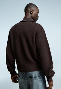 Mörkbrun stickad tröja med lös passform, som har en krage och ribbade ärmslut. Bärs tillsammans med ljusgrå denimbyxor som visar textur och design.