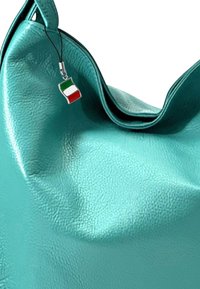 Sac en cuir turquoise à texture galet, avec une forme douce et flasque, comprenant un petit charm en forme de drapeau italien attaché à la sangle.