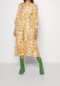 Abito midi floreale nelle tonalità crema e oro, con maniche a sbuffo e vita strizzata. Abbinato a stivali platform in suede verde.