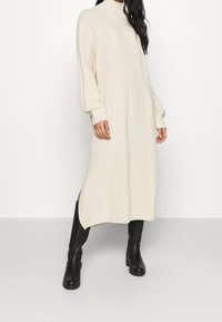 Abito midi in maglia color crema con collo alto, spalle scese e spacchi laterali, abbinato a stivali neri con tacco alti fino al ginocchio.