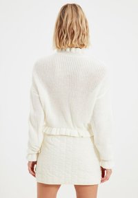 Žena s blond vlasy, oblečená v bílém silném pleteném svetru a bílé texturované sukni, viděná zezadu na jednobarevném pozadí.