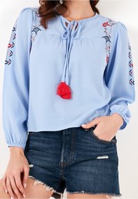 Blouse bleu clair à manches longues, fermeture avec lien au col, et détail de pompon rouge. Présente une broderie florale colorée sur les manches. Portée avec un short sombre.