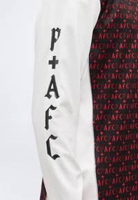 Manica bianca con scritte nere in stile gotico "p+AFC" accanto a tessuto nero decorato con motivi rossi "AFC" e segni più.