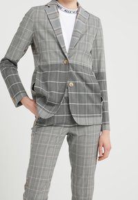 Blazer grigio a quadri con rever a punta, due bottoni e tasche laterali, abbinato a pantaloni a quadri coordinati. Tessuto liscio, vestibilità sartoriale.