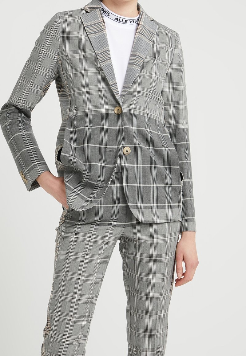 Blazer grigio a quadri con rever a punta, due bottoni e tasche laterali, abbinato a pantaloni a quadri coordinati. Tessuto liscio, vestibilità sartoriale.