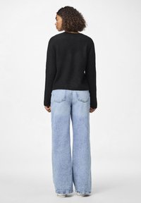 Suéter preto de malha com mangas longas, combinado com jeans de perna larga azul claro, apresentando fecho de botão e zíper e bolsos traseiros.