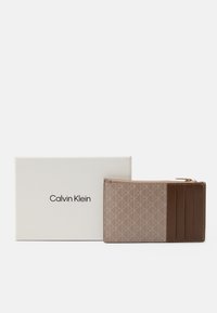 Porte-cartes Calvin Klein en cuir tan et marron avec un motif géométrique. Comprend plusieurs emplacements pour cartes et une poche zippée. Inclus une boîte blanche.