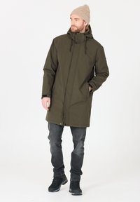 Parka impermeabile verde oliva con cappuccio, dotata di cerniera frontale, chiusura a pressione e due tasche laterali. Indossato sopra jeans scuri e scarponi da trekking.