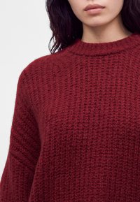 Femme portant un pull tricoté texturé rouge foncé avec un col montant, montrant uniquement la partie inférieure de son visage et le haut de son torse.