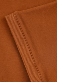 Tessuto arancione bruciato con una texture a righe, che mostra pieghe e dettagli delle cuciture. Il materiale è morbido e si allunga leggermente.